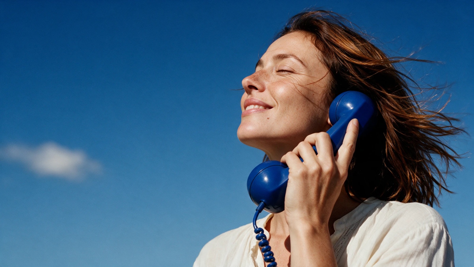 Mulher sorrindo de olhos fechados ao telefone azul, contra o céu